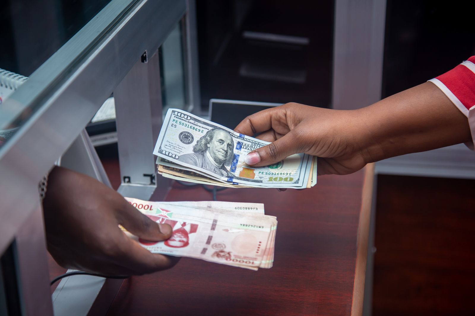 Mãos trocando cédulas de moedas diferentes sobre um balcão de banco, com notas de dólar e de real lado a lado.